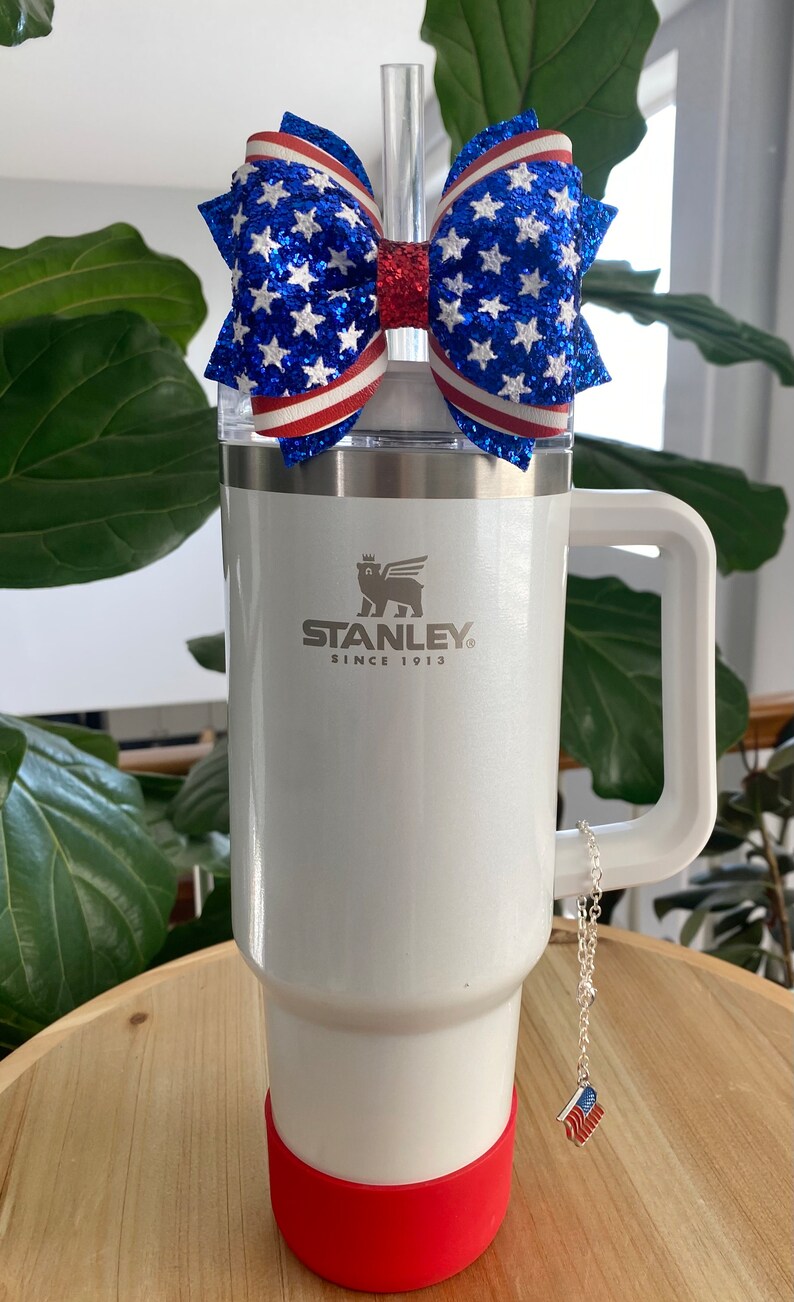 May include: White Stanley travel mug with a red, white, and blue glitter bow on top. The mug has a handle and a red silicone base. The mug has the Stanley logo and the text "Since 1913". A silver chain with a small American flag charm hangs from the handle.
