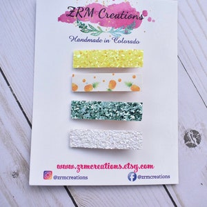 May include: Set of four hair clips with different designs. The clips are yellow glitter, white with pineapple print, green glitter, and white glitter. The clips are displayed on a white card with the text "ZRM Creations" and "Handmade in Colorado".