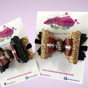 May include: Two hair bows with black and gold glitter and a brown football design. The bows are on white cards with the text "ZRM Creations" and "Handmade in Col" and the website address "www.zrmcreations.etsy.com".