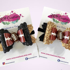 May include: Two handmade hair bows with a football theme. One bow is black with gold glitter and the other is gold with black glitter. Both bows feature a brown football with white stitching.