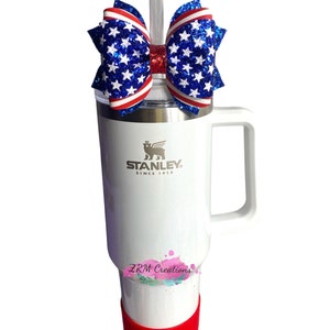 May include: A white Stanley travel mug with a red lid and a red, white, and blue glitter bow on top. The mug has the Stanley logo and the words "Since 1913" on it. The bow has a clear straw sticking out of it. The mug also has the words "ZRM Creations" on it.