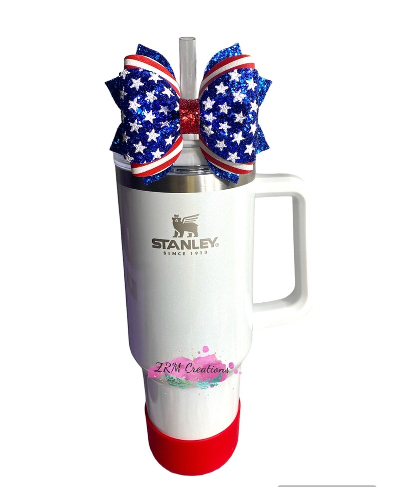 May include: A white Stanley travel mug with a red lid and a red, white, and blue glitter bow on top. The mug has the Stanley logo and the words "Since 1913" on it. The bow has a clear straw sticking out of it. The mug also has the words "ZRM Creations" on it.