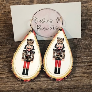 May include: A pair of dangle earrings featuring a nutcracker design with a leopard print pattern. The earrings are white with a gold trim and red and black plaid accents. The earrings are labeled "Rubies & Biscuits".