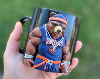 Puede incluir: Taza de cerámica negra con un oso esculpido en 3D que lleva una camiseta azul y naranja con la palabra "Bears" en ella. El oso sostiene un balón de fútbol.