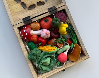 Puede incluir: Una caja de madera con un diseño tallado en la tapa está abierta, revelando una colección de verduras y champiñones de arcilla polimérica hechos a mano y de colores vivos. La caja está llena de material parecido a la paja.