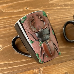 Peut inclure: Mug en céramique noire avec un motif 3D de coléoptère cerf sur fond brun et vert.
