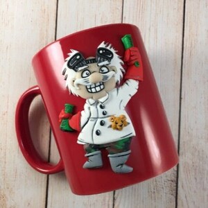 May include: Red ceramic mug with a 3D sculpted image of a scientist wearing a white lab coat and holding a test tube. The scientist has a white beard and mustache and is wearing a black hat.