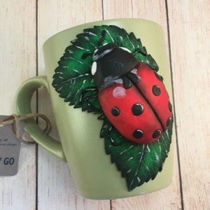 Pode incluir: Uma caneca de cerâmica verde com um design de joaninha 3D. A joaninha é vermelha com manchas pretas e está sentada em uma folha verde.