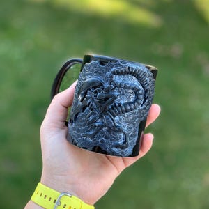 Peut inclure: Une tasse en c&eacute;ramique noire avec un motif d'extraterrestre sculpt&eacute; en 3D. La tasse est tenue dans une main avec un bracelet jaune.