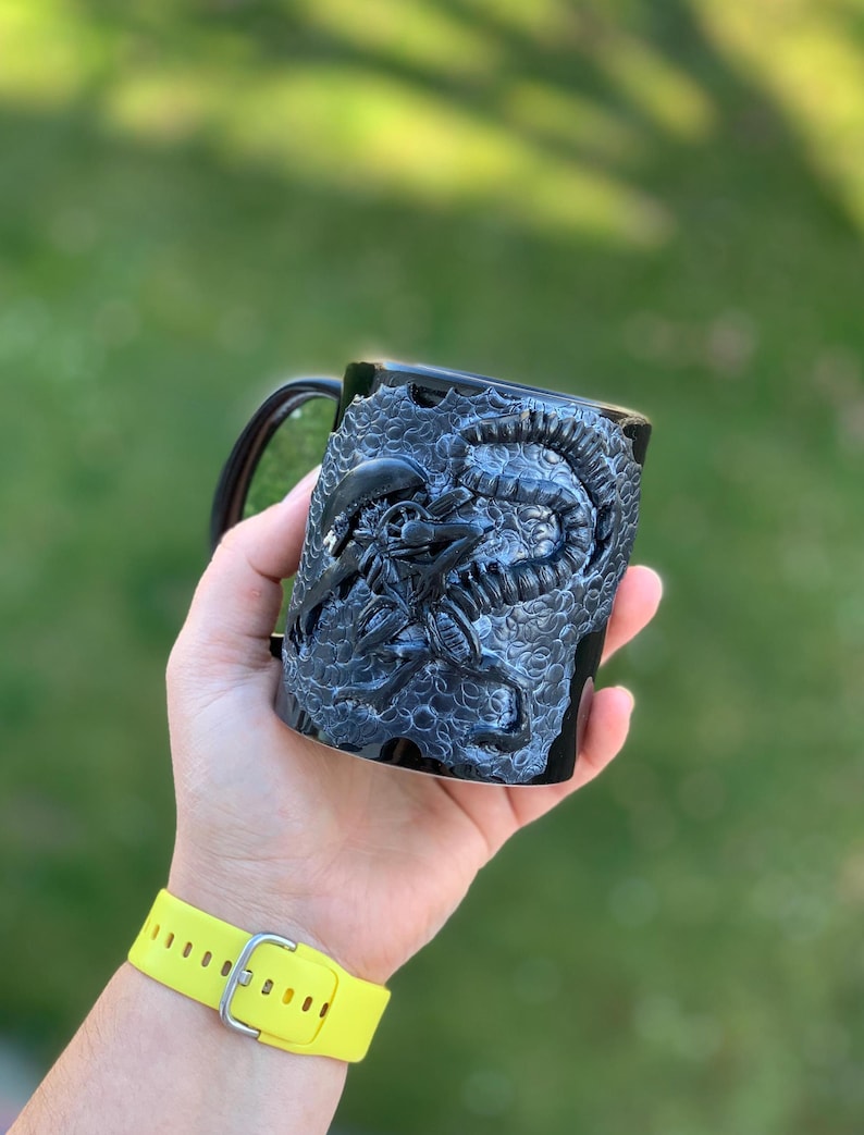 Peut inclure: Une tasse en c&eacute;ramique noire avec un motif d'extraterrestre sculpt&eacute; en 3D. La tasse est tenue dans une main avec un bracelet jaune.