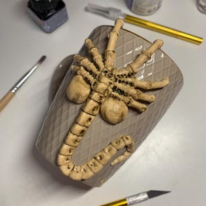 May include: A beige ceramic mug with a raised, detailed, cream-colored alien facehugger design. The mug has a diamond pattern and is surrounded by art supplies, including paint bottles and a craft knife.