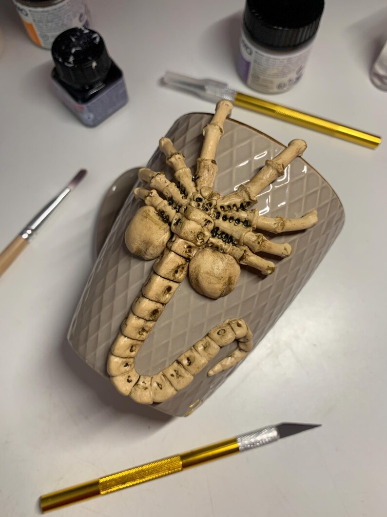 May include: A beige ceramic mug with a raised, detailed, cream-colored alien facehugger design. The mug has a diamond pattern and is surrounded by art supplies, including paint bottles and a craft knife.