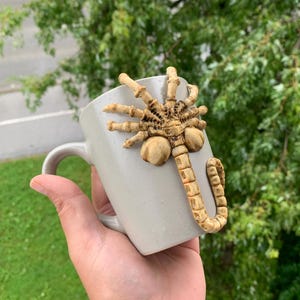 May include: A light gray ceramic mug with a sculpted, tan-colored facehugger design attached to the side. The mug has a curved handle and is held against a blurred green background.