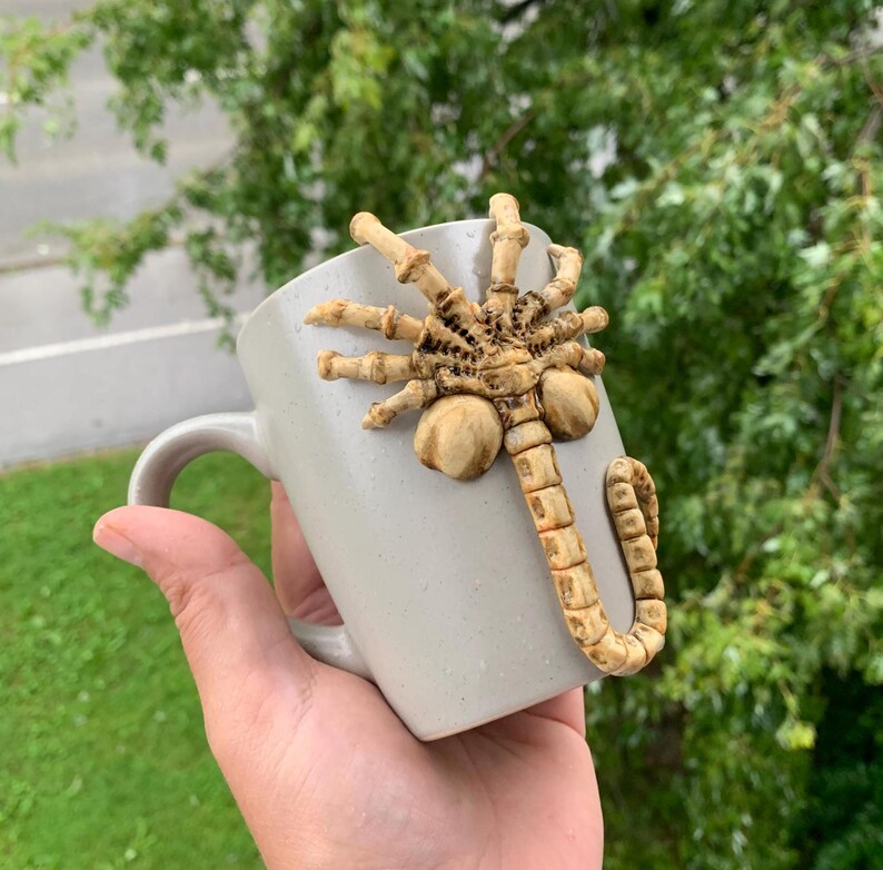 May include: A light gray ceramic mug with a sculpted, tan-colored facehugger design attached to the side. The mug has a curved handle and is held against a blurred green background.