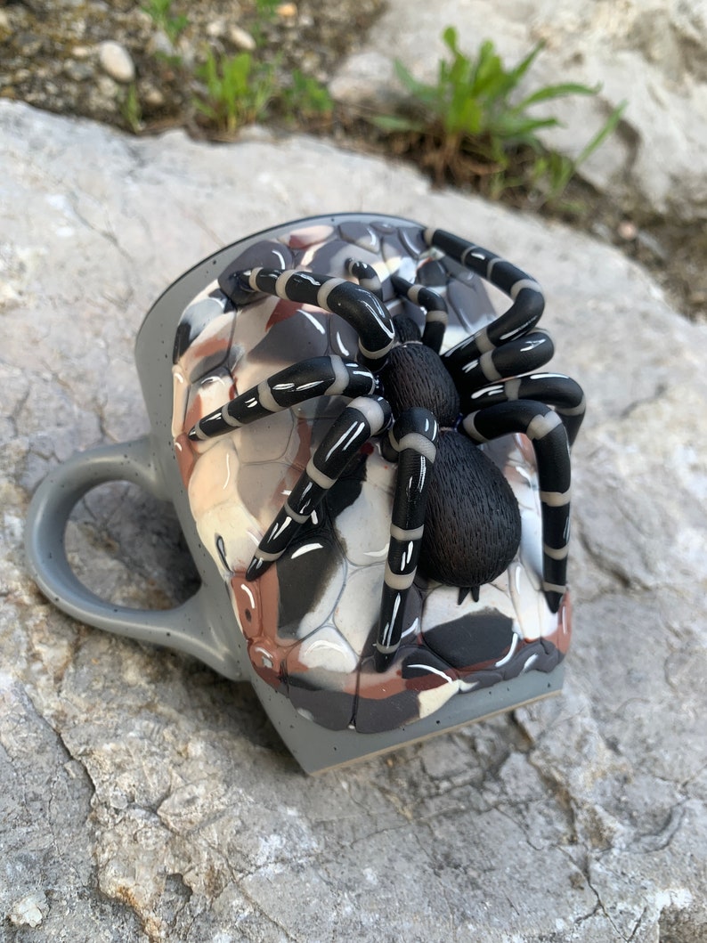 Puede incluir: Taza de cer&aacute;mica gris con una gran escultura de ara&ntilde;a. La ara&ntilde;a es negra con rayas blancas en las patas y un cuerpo peludo. La taza tiene un patr&oacute;n de camuflaje en tonos grises, marrones y blancos.