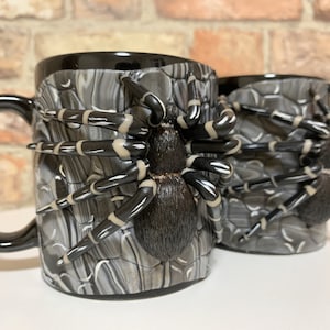 Puede incluir: Dos tazas de caf&eacute; de cer&aacute;mica con asa y borde negros. Las tazas est&aacute;n decoradas con un dise&ntilde;o de ara&ntilde;a tar&aacute;ntula en 3D. La ara&ntilde;a es negra con rayas blancas y beige en las patas. El cuerpo de la taza tiene una textura de piedra gris.