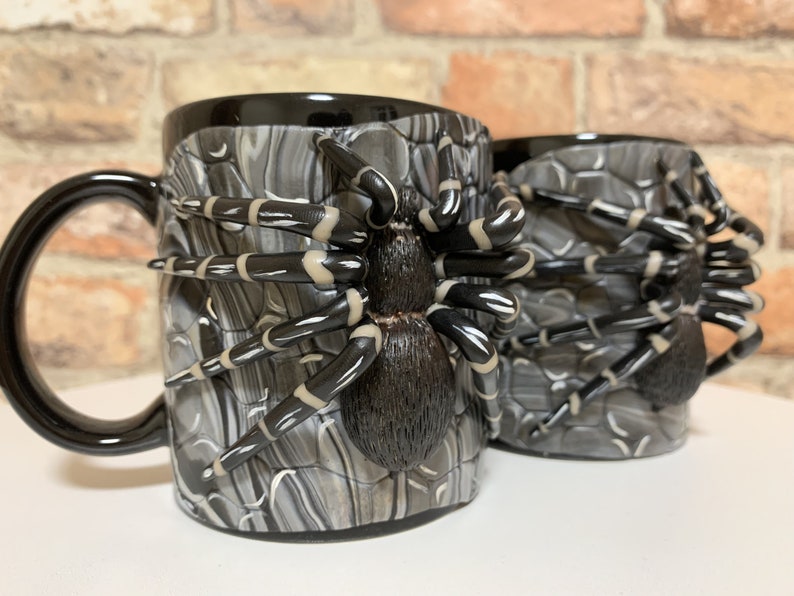 Puede incluir: Dos tazas de caf&eacute; de cer&aacute;mica con asa y borde negros. Las tazas est&aacute;n decoradas con un dise&ntilde;o de ara&ntilde;a tar&aacute;ntula en 3D. La ara&ntilde;a es negra con rayas blancas y beige en las patas. El cuerpo de la taza tiene una textura de piedra gris.