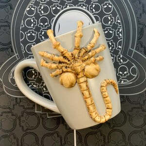 May include: A light gray ceramic mug with a detailed, tan-colored Xenomorph facehugger design. The mug is set against a black background with a skull pattern. The mug has a curved handle.