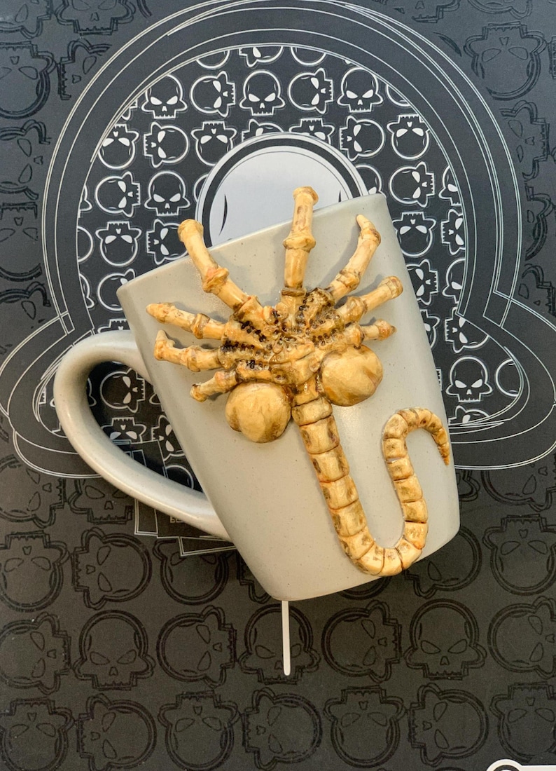 May include: A light gray ceramic mug with a detailed, tan-colored Xenomorph facehugger design. The mug is set against a black background with a skull pattern. The mug has a curved handle.