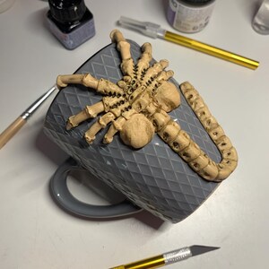 May include: A gray ceramic mug with a raised diamond pattern, adorned with a beige, detailed spider sculpture. The mug is resting on its side, with a gold-handled craft knife and other art supplies visible in the background.