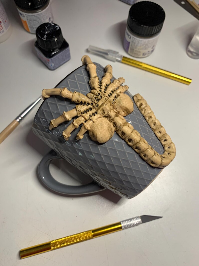 May include: A gray ceramic mug with a raised diamond pattern, adorned with a beige, detailed spider sculpture. The mug is resting on its side, with a gold-handled craft knife and other art supplies visible in the background.