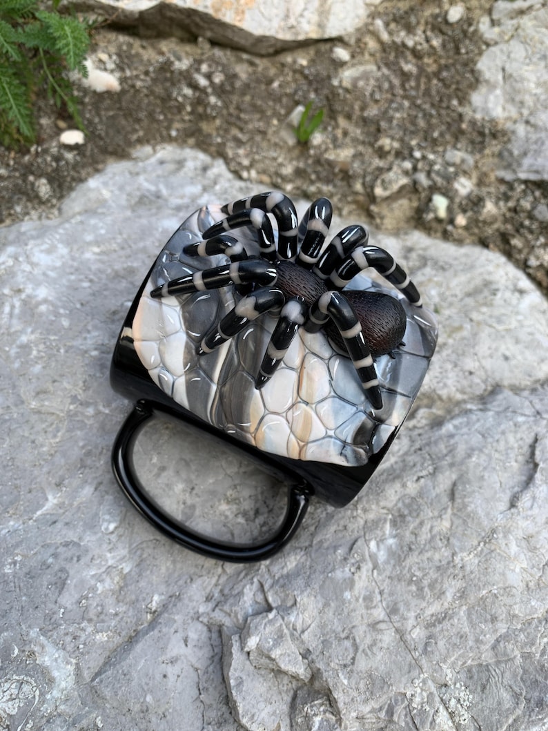 Puede incluir: Taza de caf&eacute; negra con una ara&ntilde;a decorativa en la parte superior. La ara&ntilde;a tiene patas a rayas blancas y negras y un cuerpo marr&oacute;n. La superficie de la taza tiene un patr&oacute;n gris y blanco.