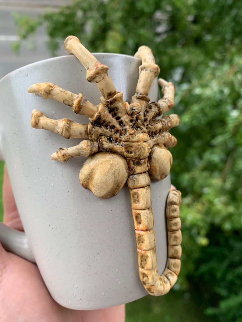 May include: A gray ceramic mug with a detailed, beige-colored alien facehugger design attached to the side. The facehugger has multiple segmented legs and a long, curved tail. The mug has a simple handle and a speckled texture.