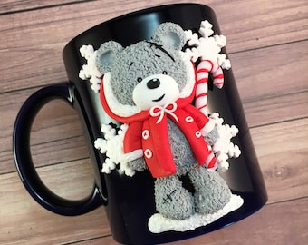 Puede incluir: Una taza de cerámica azul con un oso de peluche gris que lleva un abrigo rojo y sostiene un bastón de caramelo. El oso está rodeado de copos de nieve blancos.