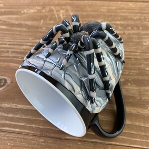 Puede incluir: Taza de caf&eacute; negra con una gran escultura de ara&ntilde;a detallada en el lateral. La ara&ntilde;a tiene patas a rayas negras y blancas y un cuerpo texturizado. El interior de la taza es blanco y el asa es negra.
