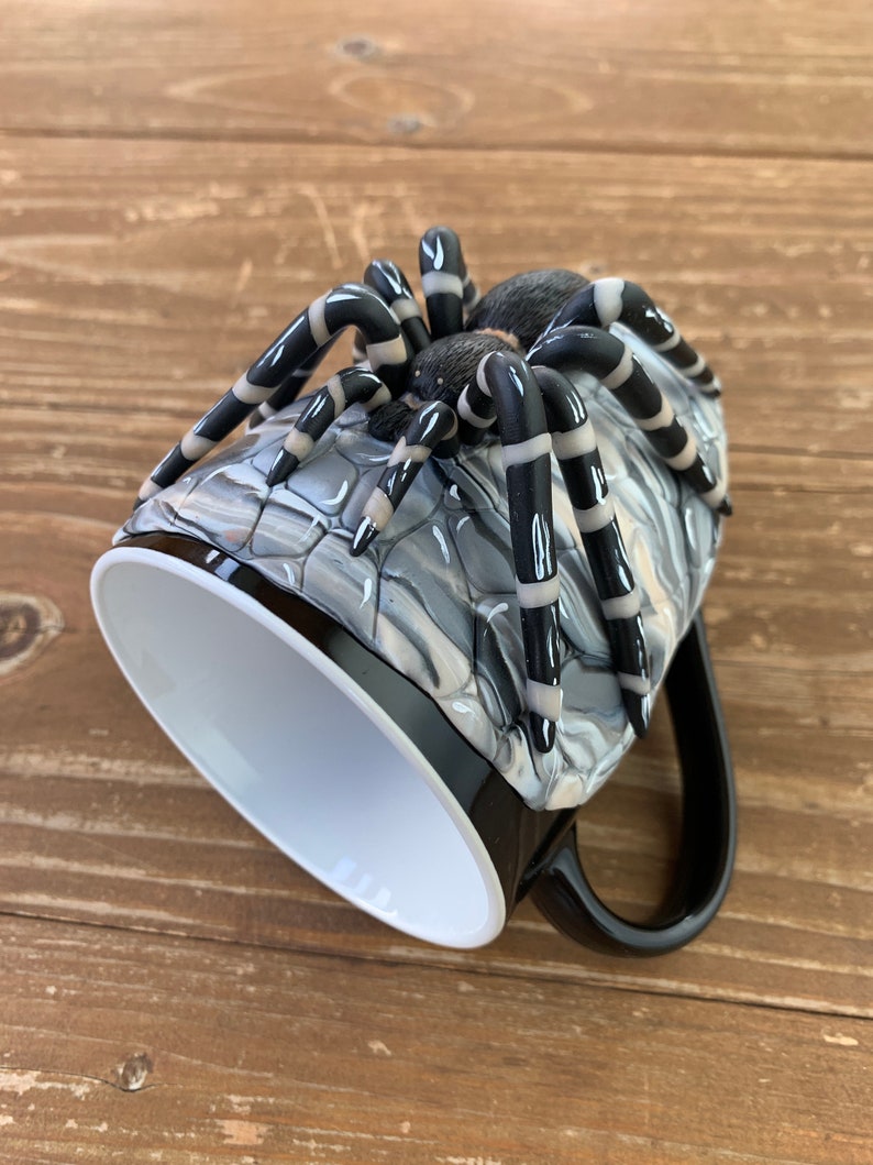 Puede incluir: Taza de caf&eacute; negra con una gran escultura de ara&ntilde;a detallada en el lateral. La ara&ntilde;a tiene patas a rayas negras y blancas y un cuerpo texturizado. El interior de la taza es blanco y el asa es negra.
