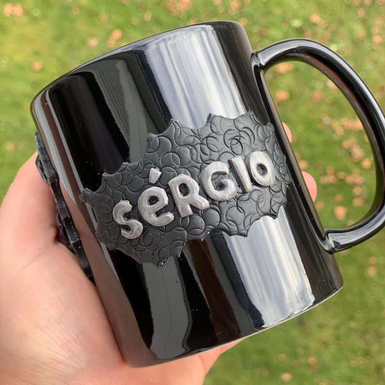 Peut inclure: Mug en c&eacute;ramique noire avec un nom en relief argent&eacute; "Sergio" sur un fond noir textur&eacute;.