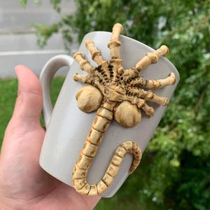 May include: A light gray ceramic mug with a sculpted, beige-colored alien facehugger design. The facehugger is attached to the mug's surface, with its legs and tail wrapping around the cup. The mug has a simple, curved handle.