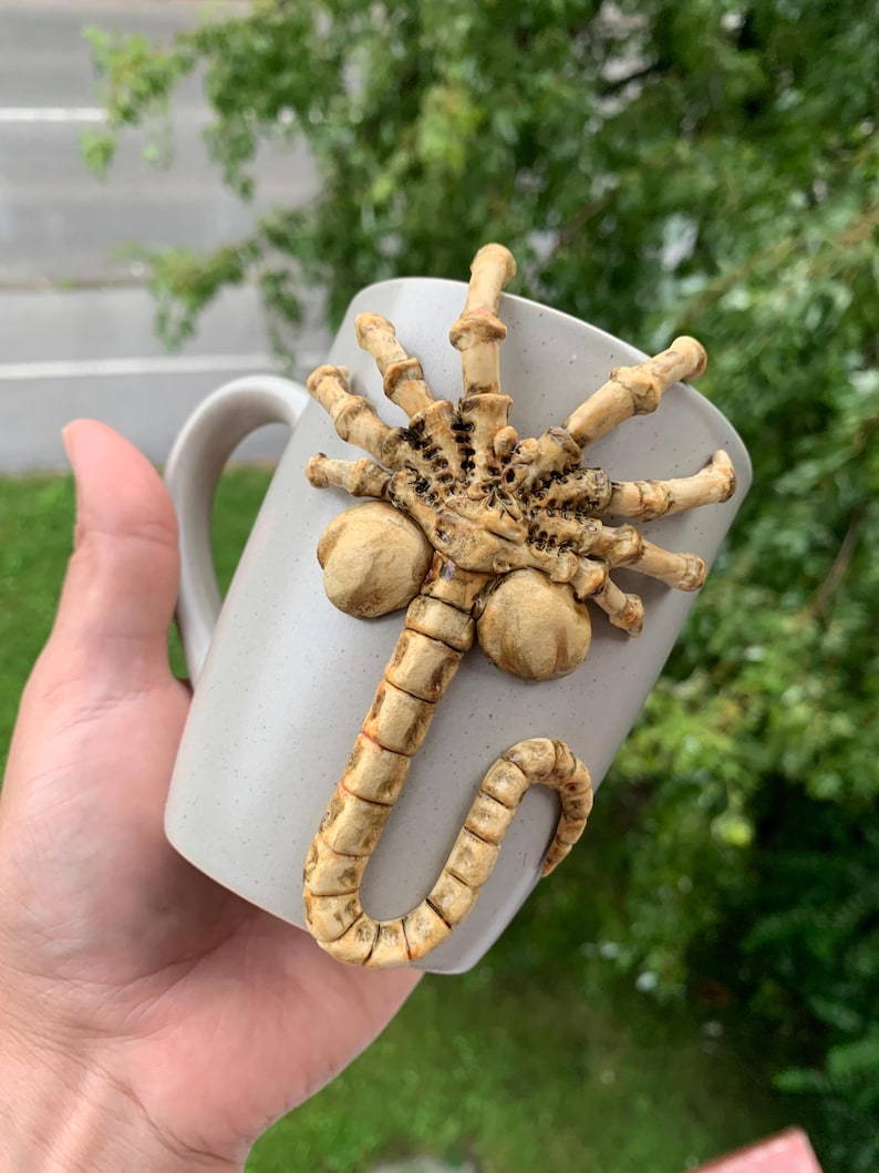 May include: A light gray ceramic mug with a sculpted, beige-colored alien facehugger design. The facehugger is attached to the mug's surface, with its legs and tail wrapping around the cup. The mug has a simple, curved handle.