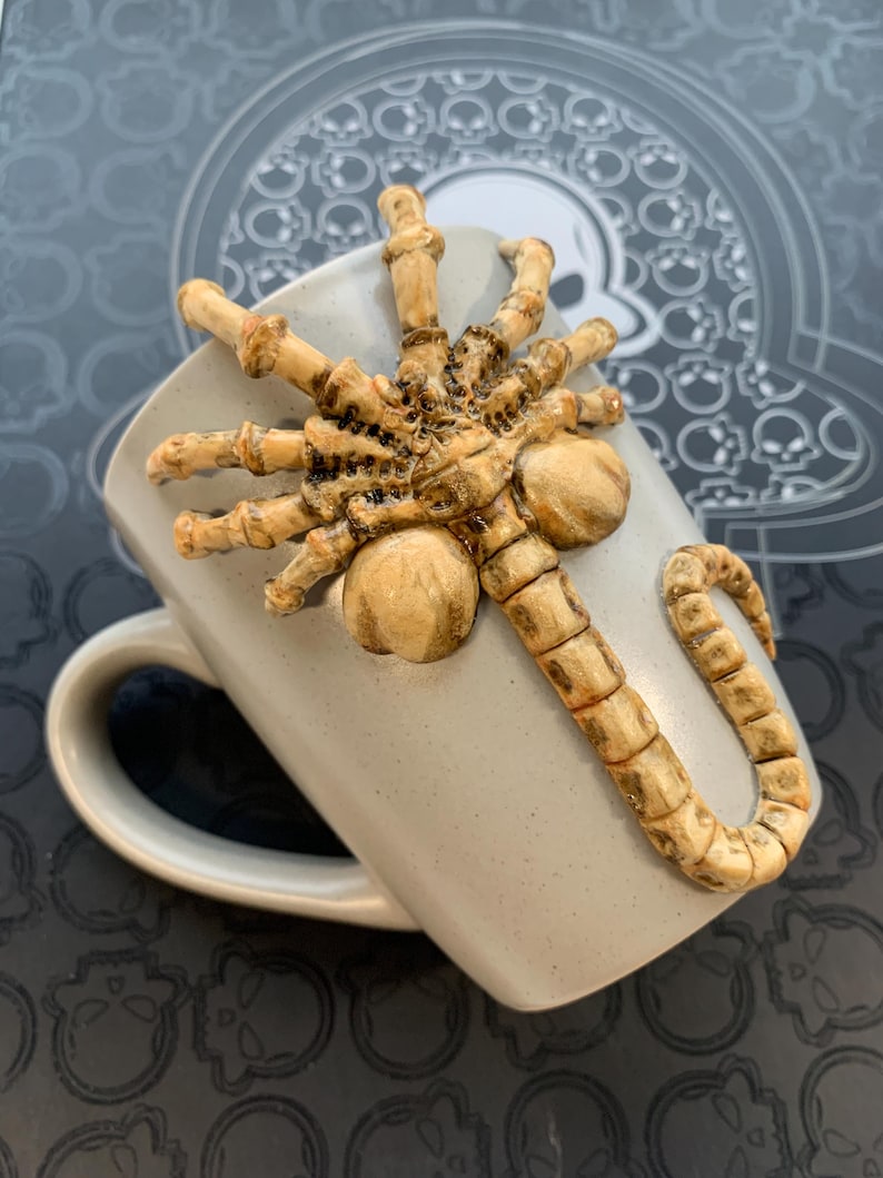 May include: A gray ceramic mug with a detailed, tan-colored alien facehugger design. The facehugger is sculpted to appear as if it is crawling across the mug's surface. The mug has a curved handle and a speckled finish.