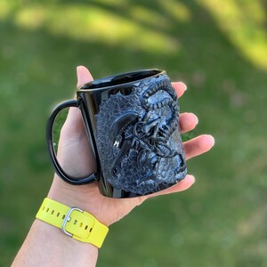 Peut inclure: Mug en c&eacute;ramique noire avec un motif sculpt&eacute; en 3D d'une cr&eacute;ature extraterrestre. La tasse a une poign&eacute;e noire et une surface textur&eacute;e.