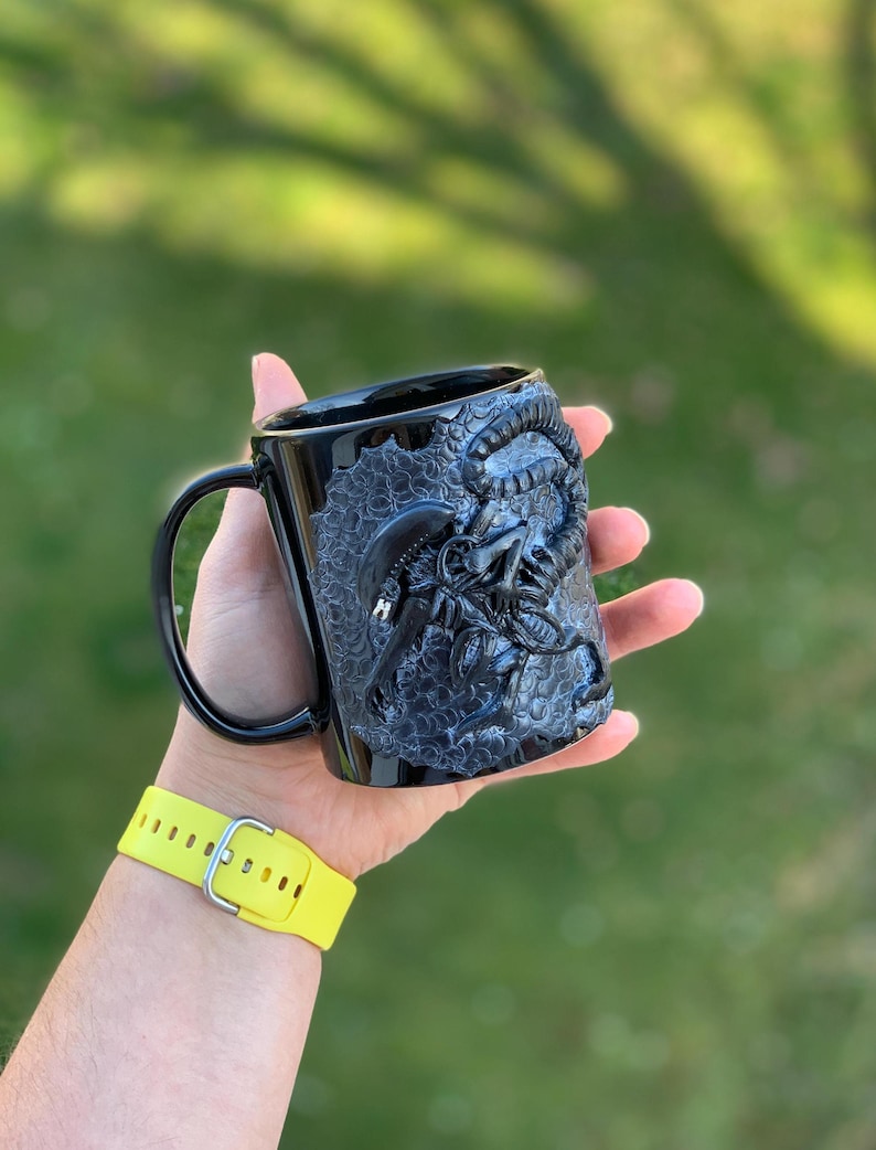 Peut inclure: Mug en c&eacute;ramique noire avec un motif sculpt&eacute; en 3D d'une cr&eacute;ature extraterrestre. La tasse a une poign&eacute;e noire et une surface textur&eacute;e.