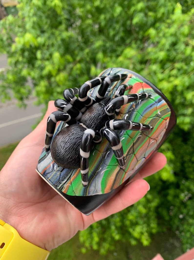 Puede incluir: Una figura de ara&ntilde;a en blanco y negro con patas a rayas descansa sobre una base colorida y estampada. La ara&ntilde;a tiene un cuerpo texturizado y es el punto focal de la imagen. La base presenta tonos verdes, naranjas y azules.