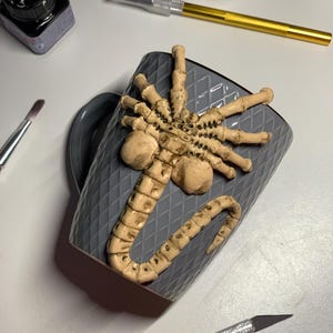 May include: A gray ceramic mug with a raised, textured design and a sculpted, beige-colored alien creature attached. The mug is surrounded by art supplies, including a brush, ink, and a craft knife.
