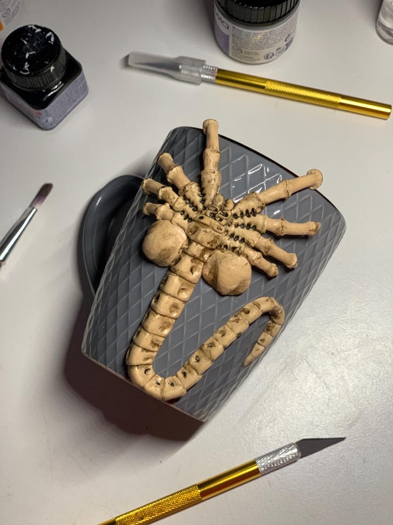 May include: A gray ceramic mug with a raised, textured design and a sculpted, beige-colored alien creature attached. The mug is surrounded by art supplies, including a brush, ink, and a craft knife.