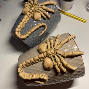 May include: Two gray ceramic mugs with a diamond pattern, each adorned with a beige Xenomorph head and tail design. The mugs are displayed on a light surface, showcasing the detailed, textured design.