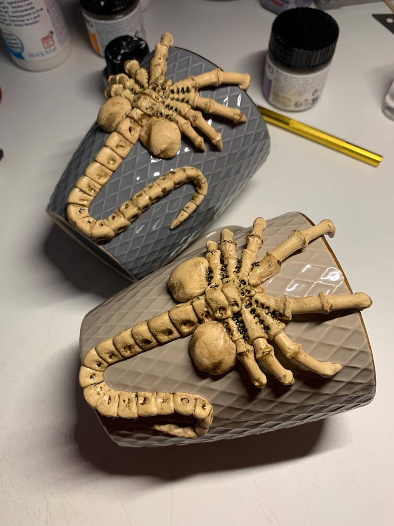May include: Two gray ceramic mugs with a diamond pattern, each adorned with a beige Xenomorph head and tail design. The mugs are displayed on a light surface, showcasing the detailed, textured design.
