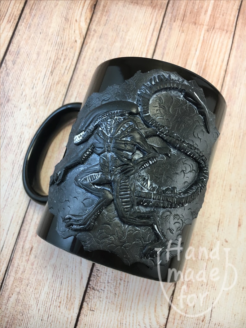 Peut inclure: Une tasse en c&eacute;ramique noire avec un motif d'extraterrestre sculpt&eacute; en 3D. L'extraterrestre est noir et a une surface textur&eacute;e et d&eacute;taill&eacute;e. La tasse a une poign&eacute;e noire et les mots "Handmade for" sont visibles sur le fond de la tasse.