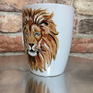 May include: White ceramic mug with a hand-painted image of a lion's face. The lion has a brown mane and a light brown face with green eyes.