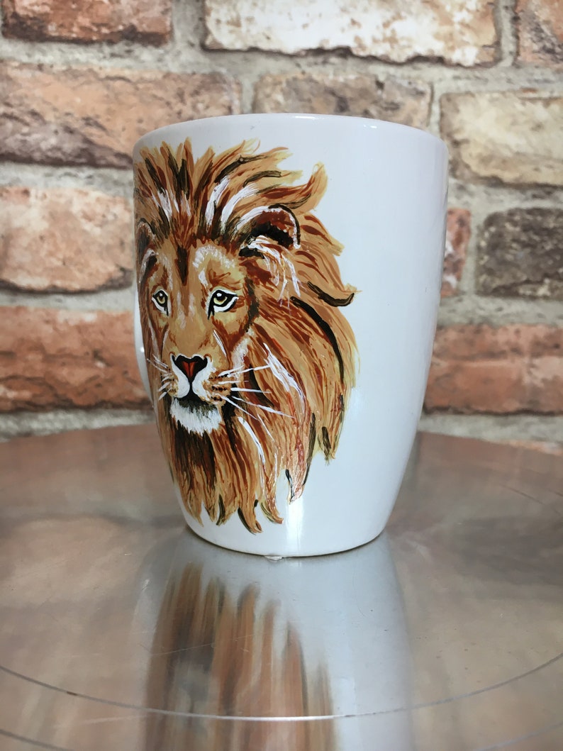 May include: White ceramic mug with a hand-painted image of a lion's face. The lion has a brown mane and a light brown face with green eyes.