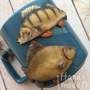 May include: A blue ceramic mug with two fish sculpted on the side. The fish are realistic and detailed, with one being a brown and gold perch and the other a striped bass. The mug has the words "Hand made for" printed on the bottom.