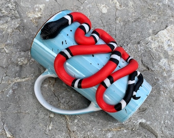 May include: A blue ceramic mug with a red, black, and white snake design wrapped around it. The snake is a king snake, a non-venomous species.