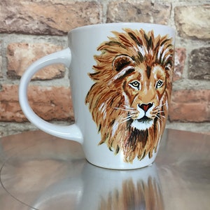 May include: White ceramic mug with a hand-painted image of a lion's face. The lion has a brown mane and a serious expression.