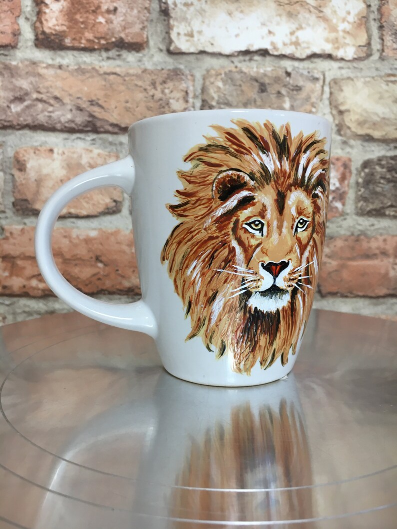May include: White ceramic mug with a hand-painted image of a lion's face. The lion has a brown mane and a serious expression.