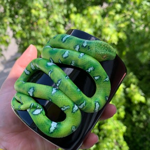 Puede incluir: Una taza de cer&aacute;mica negra con una serpiente verde esculpida enrollada alrededor. La serpiente tiene marcas blancas y verdes y una cabeza detallada. La taza se sostiene en una mano contra un fondo verde borroso.