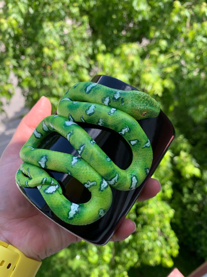 Puede incluir: Una taza de cer&aacute;mica negra con una serpiente verde esculpida enrollada alrededor. La serpiente tiene marcas blancas y verdes y una cabeza detallada. La taza se sostiene en una mano contra un fondo verde borroso.